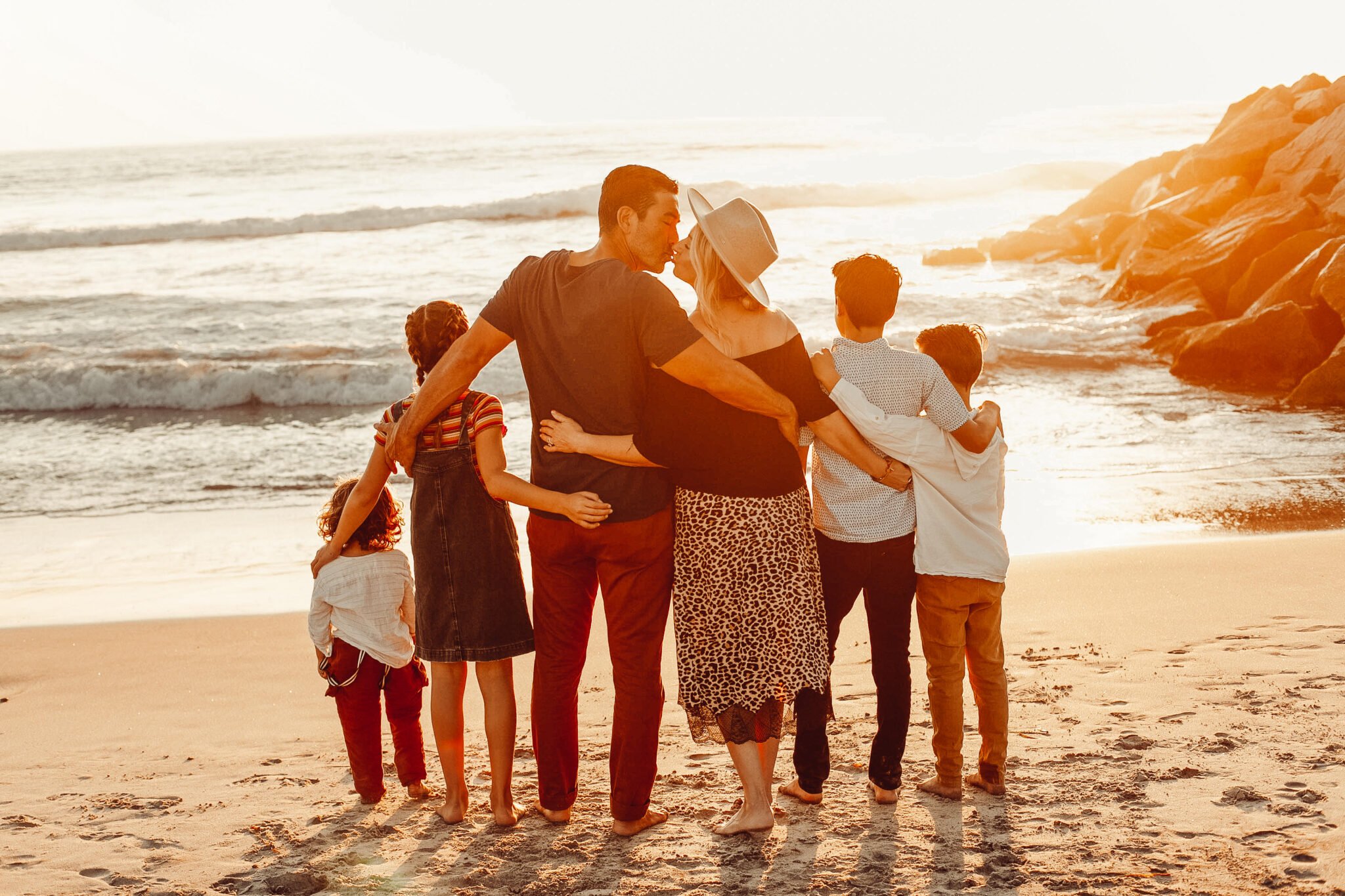 A couple kissing on the beach with their four kids standing beside them.