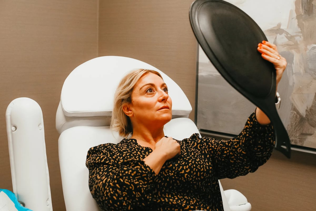 A woman lays in a white medical chair looking up at herself in a hand-held mirror after having botox injections.