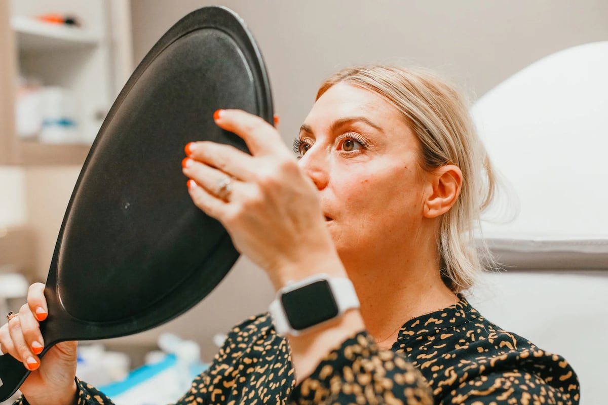 A close-up of a blonde woman holding up a small black mirror as she looks at herself.