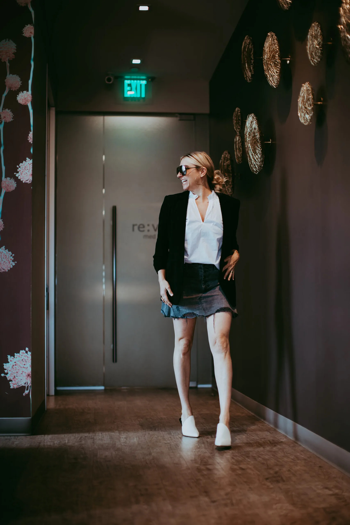 A blonde woman walks through a dark hallway of a medical spa with her sunglasses on.