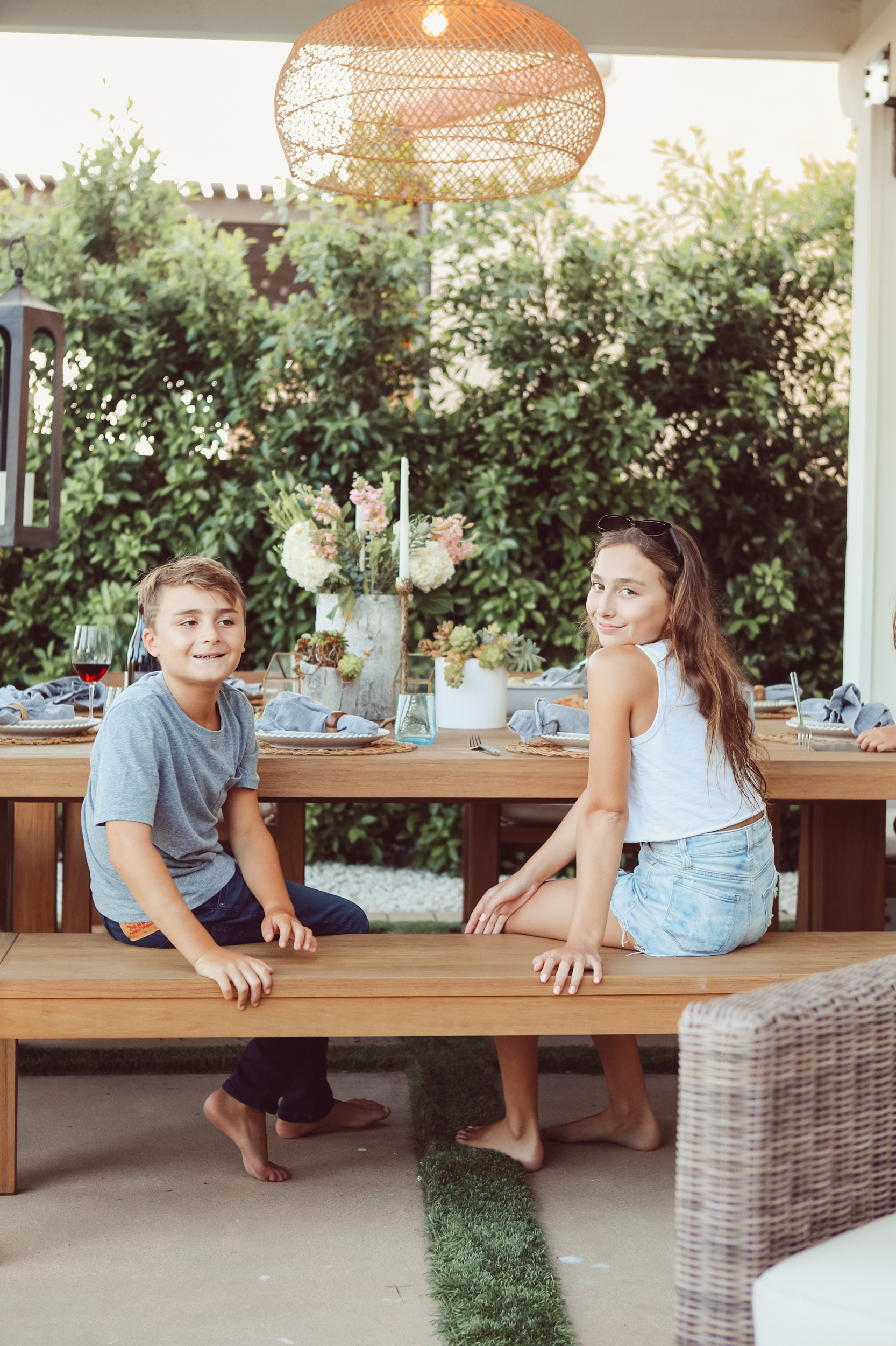 A brother and sister sit on a table in their backyard eating lunch.