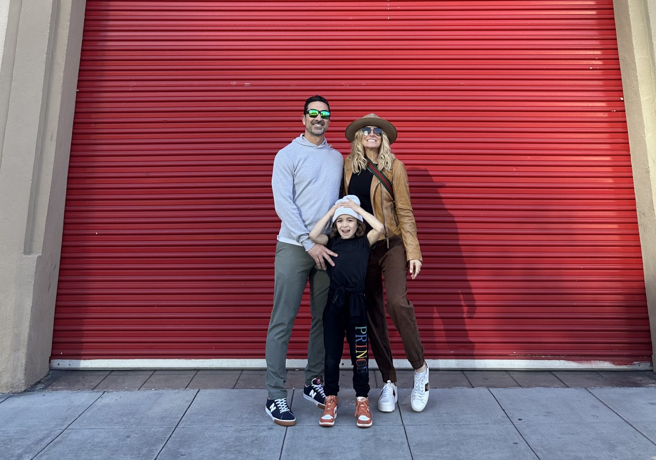 A mom and dad stand in front of a red wall with their child. 