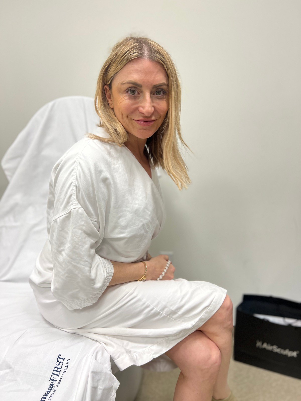 A blonde woman sits in a white robe in a doctor's office.