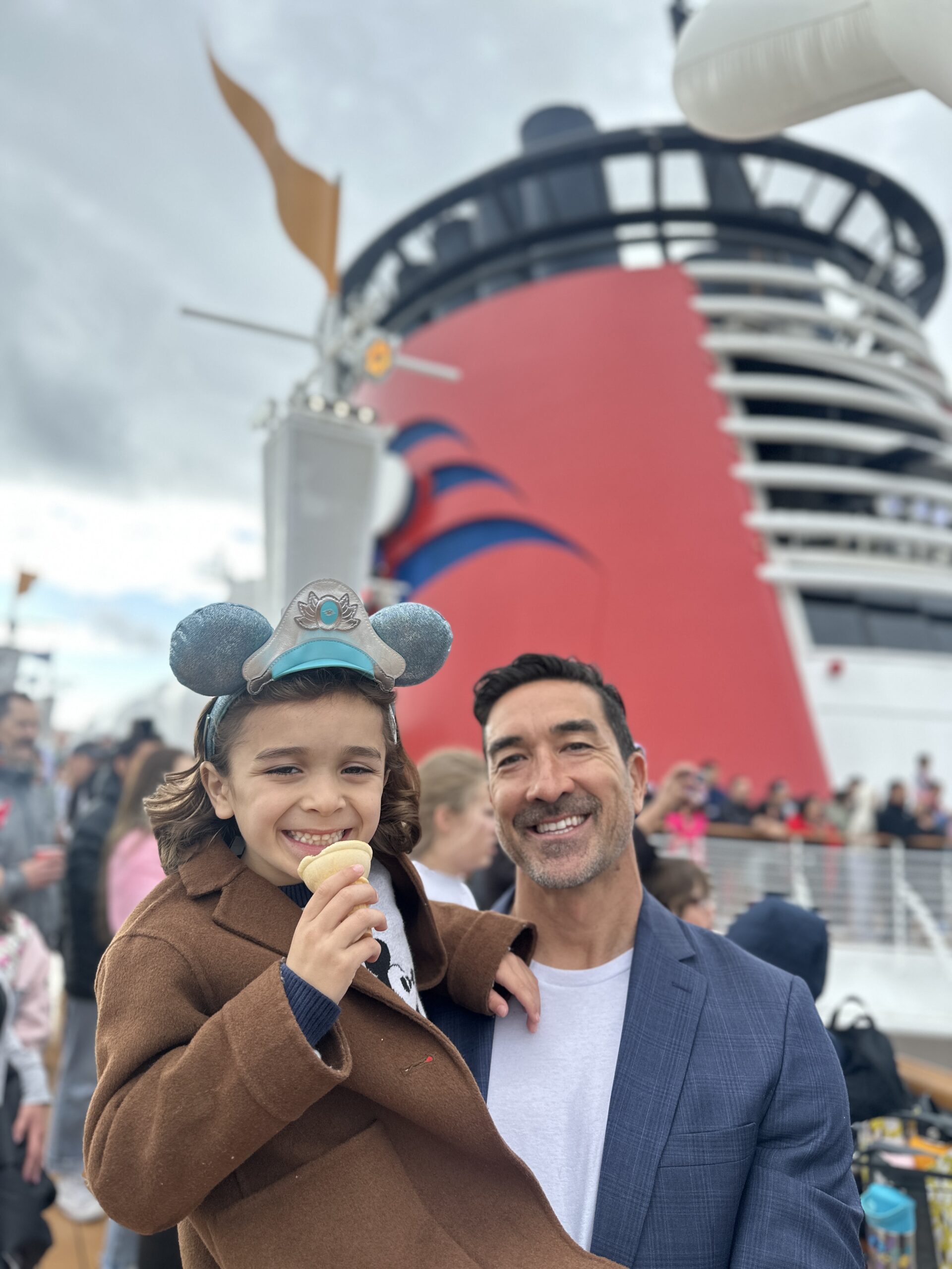 A boy in Mickey ears eats ice cream as his dad holds him in his arms.