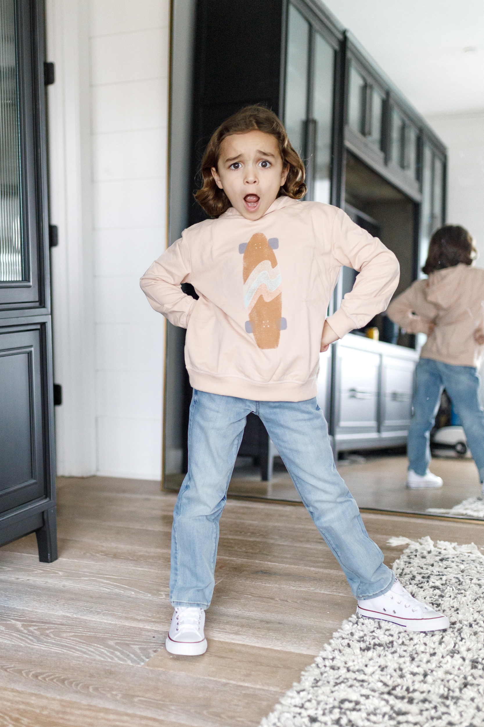 A boy strikes a fashionable pose in front of a mirror in a pink sweatshirt and jeans.