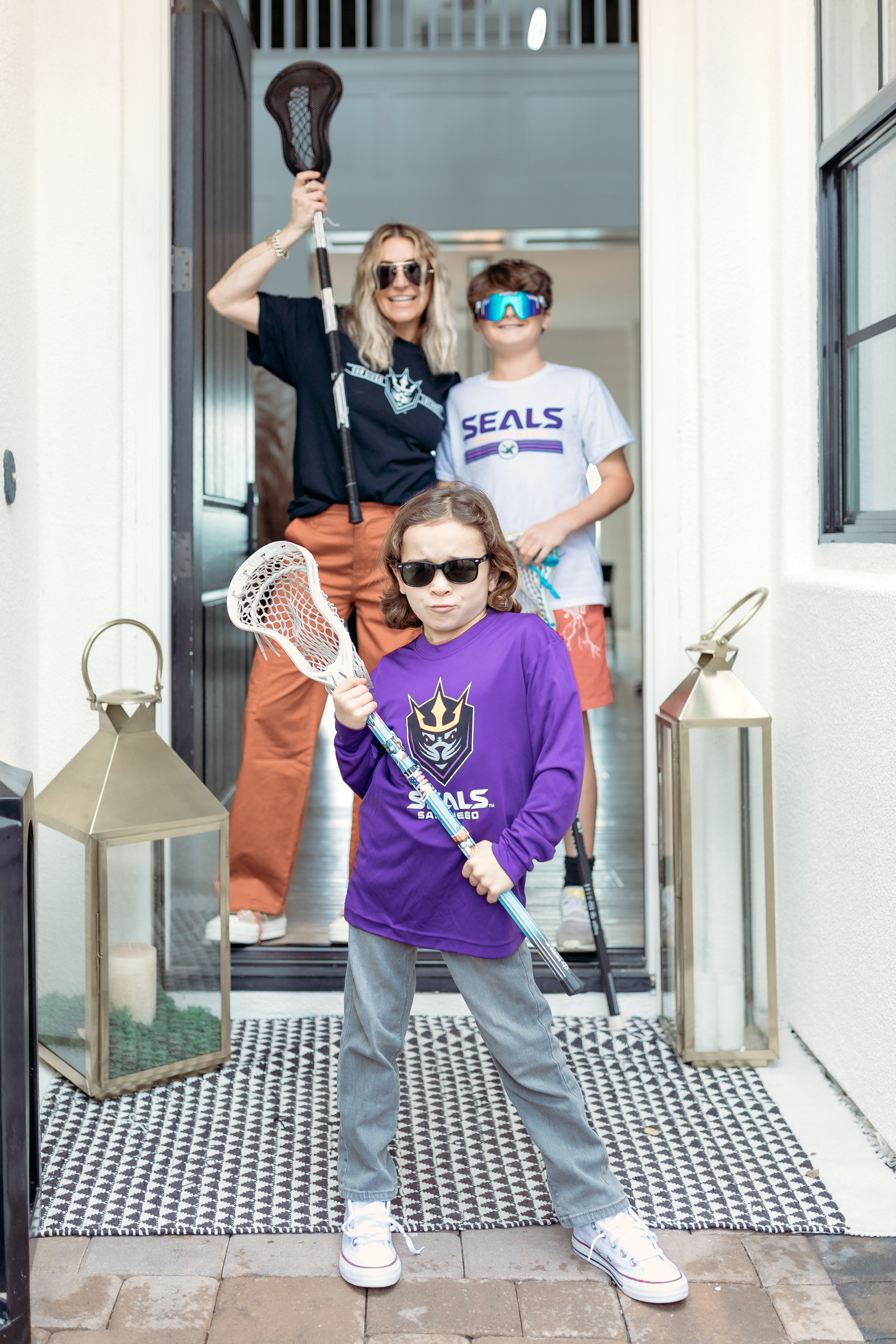 A mom and her sons walk through the front door wearing sunglasses and holding lacrosse sticks.