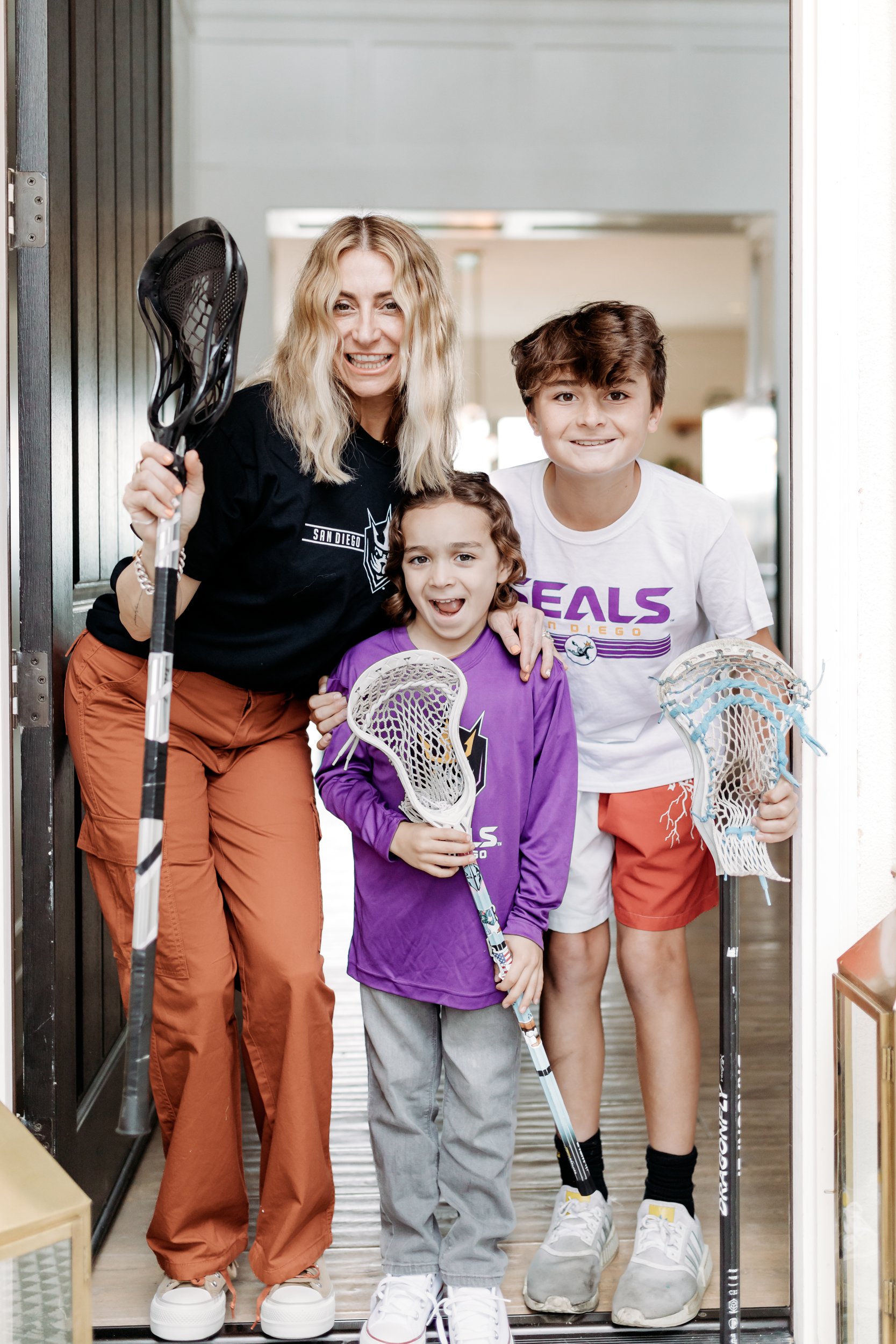 A mom and her sons pose in the doorway of their home with San Diego Seals tees on while holding lacrosse sticks. 