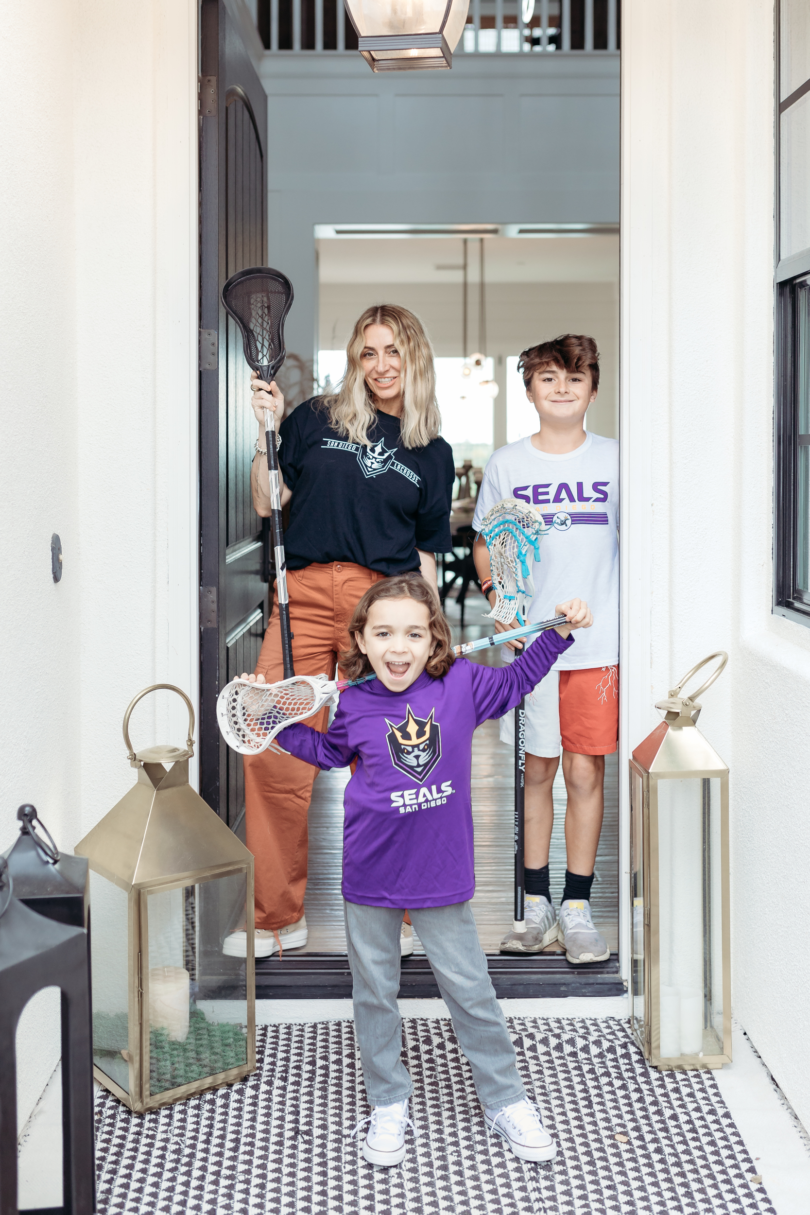A mom and her two sons stand in a doorway dressed in lacrosse fan gear and holding lacrosse sticks.