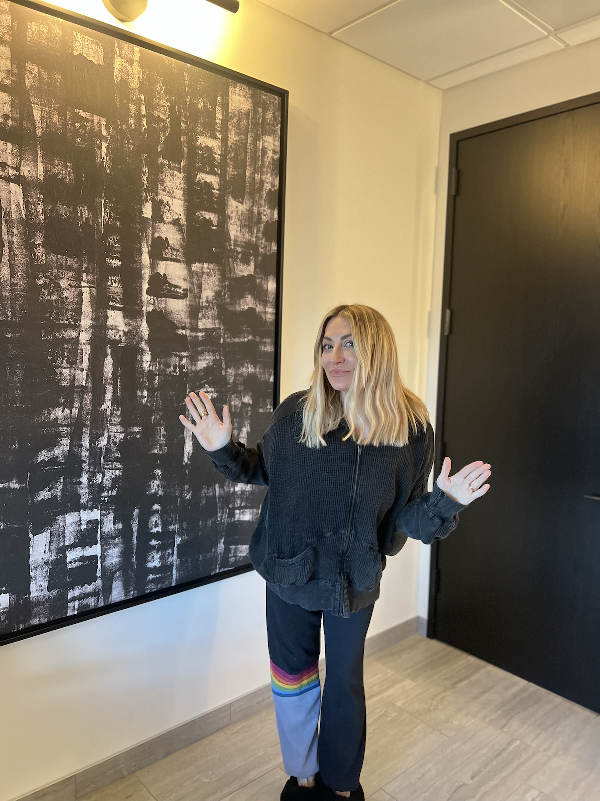 A woman stands in the corner of a doctor's office, waving at the camera.