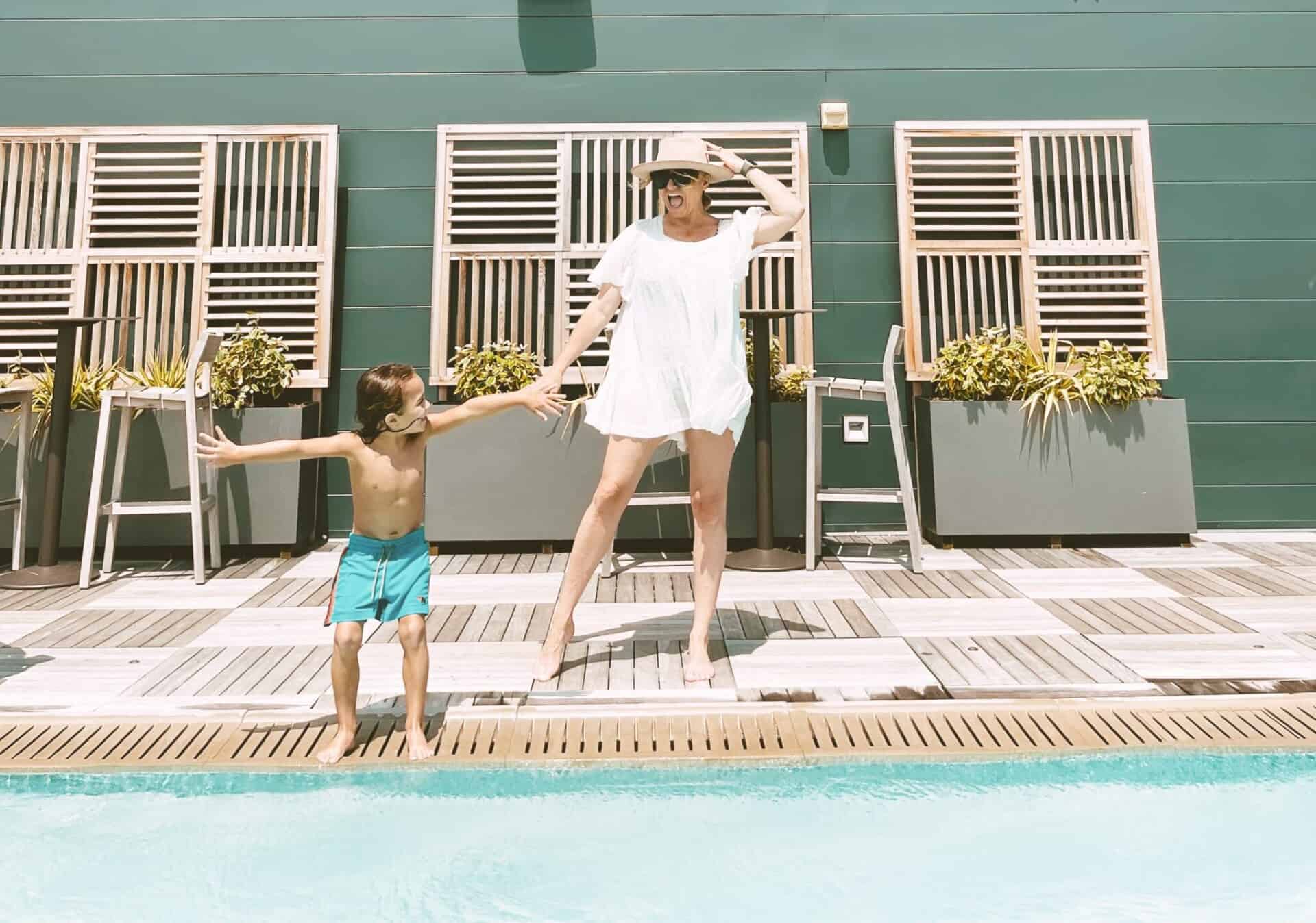 Mother and son having fun at the pool. The woman is wearing a hat and white swim coverup and the young boy is wearing blue swim trunks. He is getting ready to jump into the water.