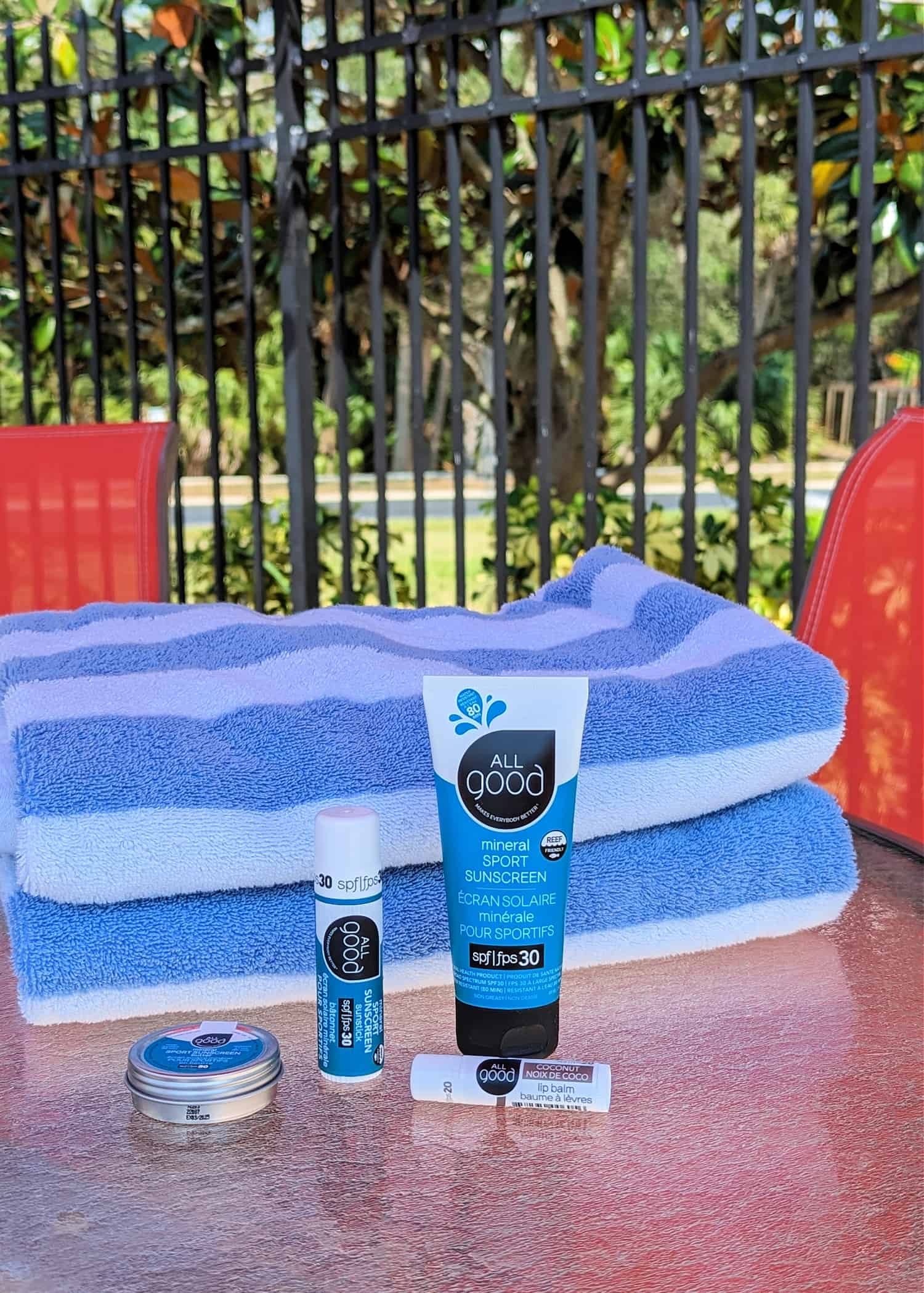 A stack of blue and white stripe beach towels on a poolside table. Several All Good Sunscreen products are sitting in front of the towels.