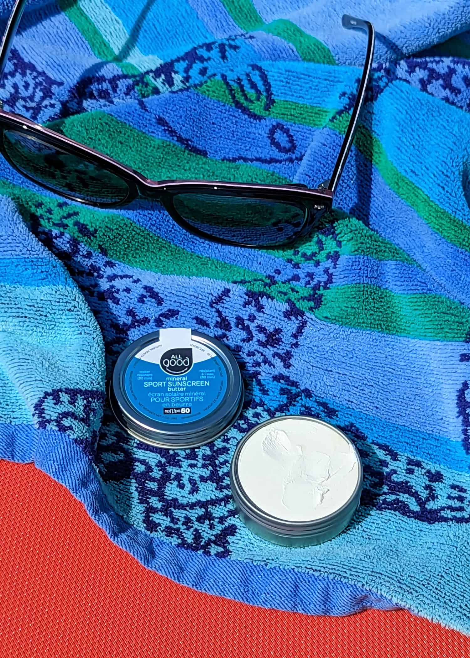 Blue beach towel on top of orange pool lounge chair. An opened tin of creamy All Good Zinc Butter sunscreen is sitting on top of the towel next to a pair of sunglasses.