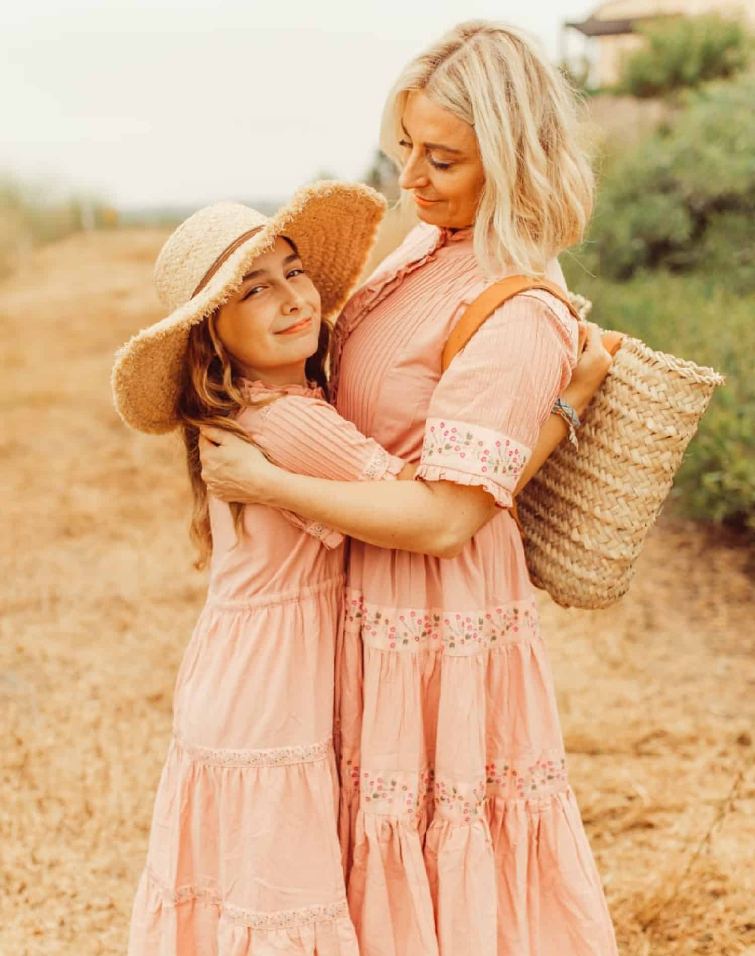 a mother and daughter smiling and hugging. They are wearing matching peach colored dresses. The girl is wearing a large brimmed straw hat and the the mother is holding a straw tote.