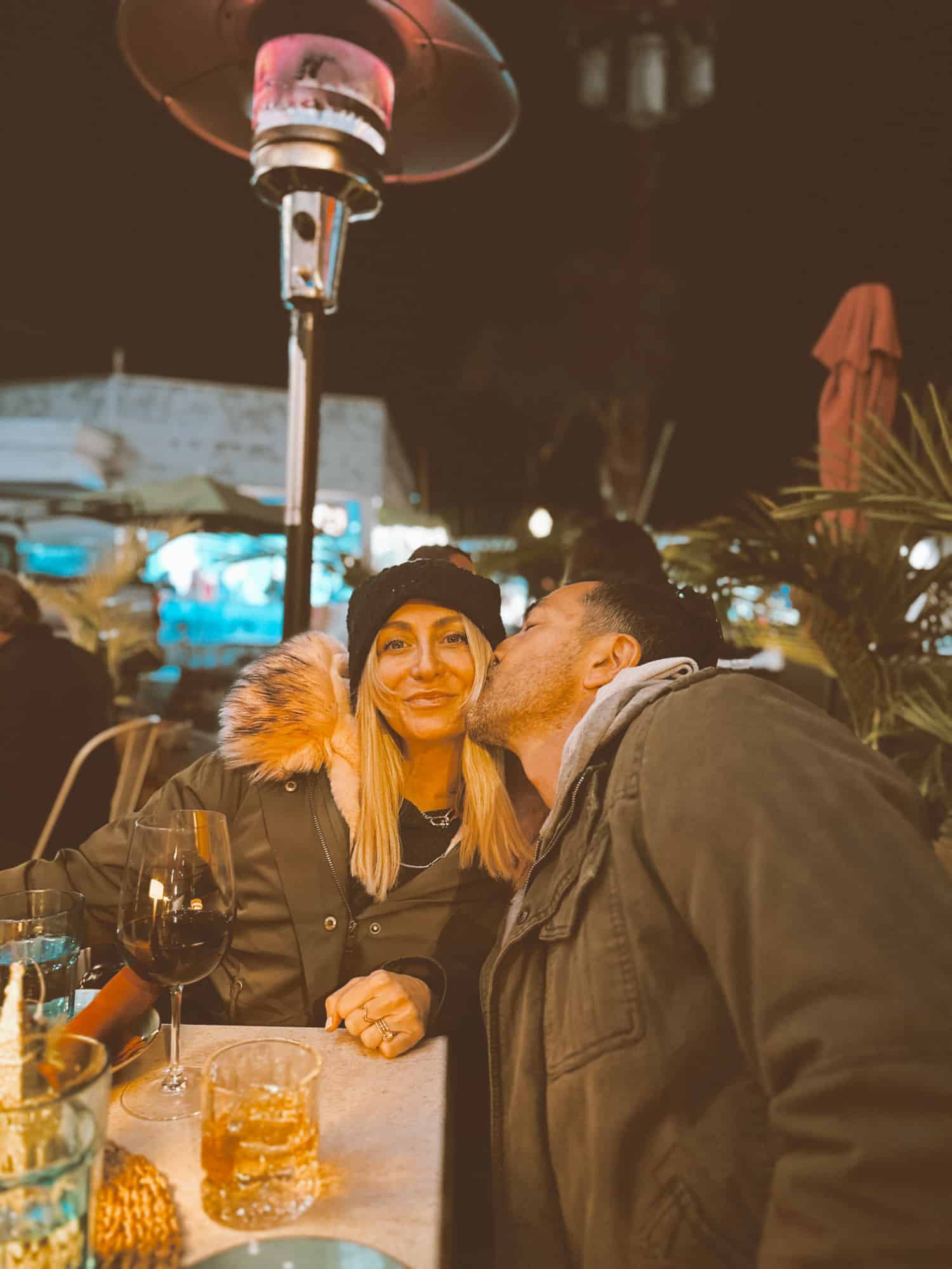 Happy parents enjoying a date night on the patio of a restaurant. The woman is smiling while being kissed on the cheek by her husband.