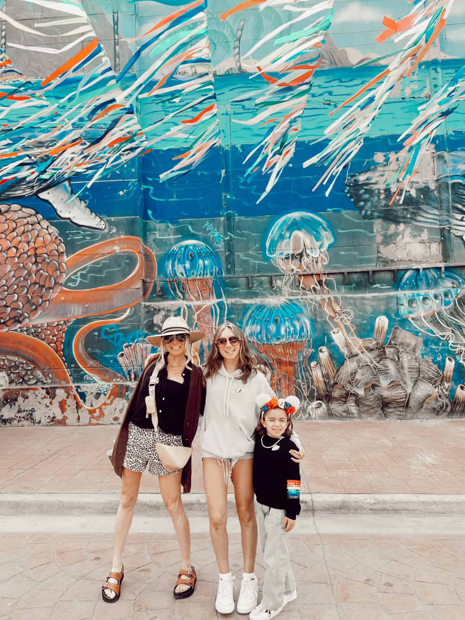 A happy mom, teen daughter and young son standing in front of a colorful ocean-themed building mural. Everyone is smiling on the sunny day. 