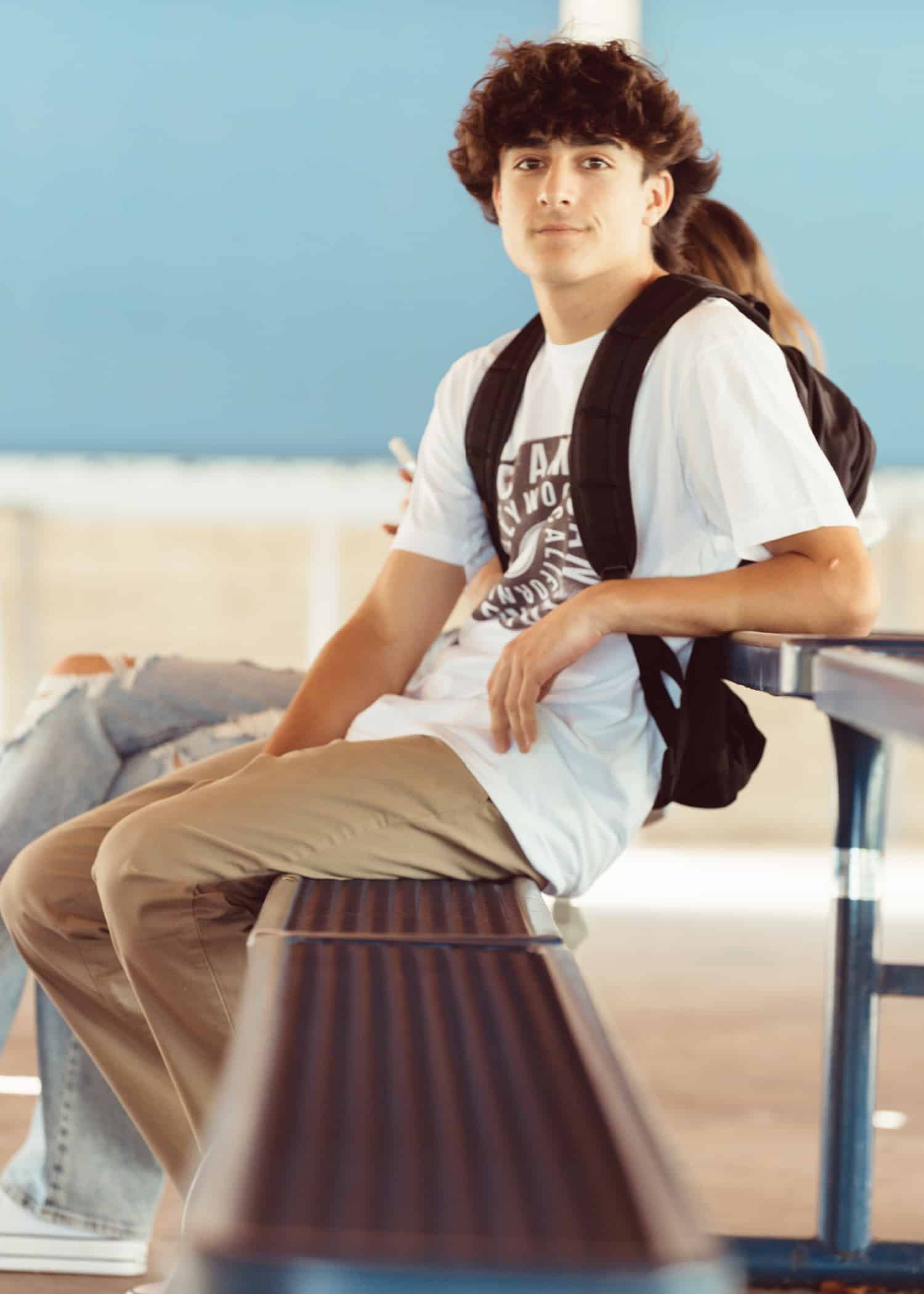 A handsome teenage boy with dark hear wearing a backpack. He is sitting on a bench. A good example of a casting profile image for a teenage actor.
