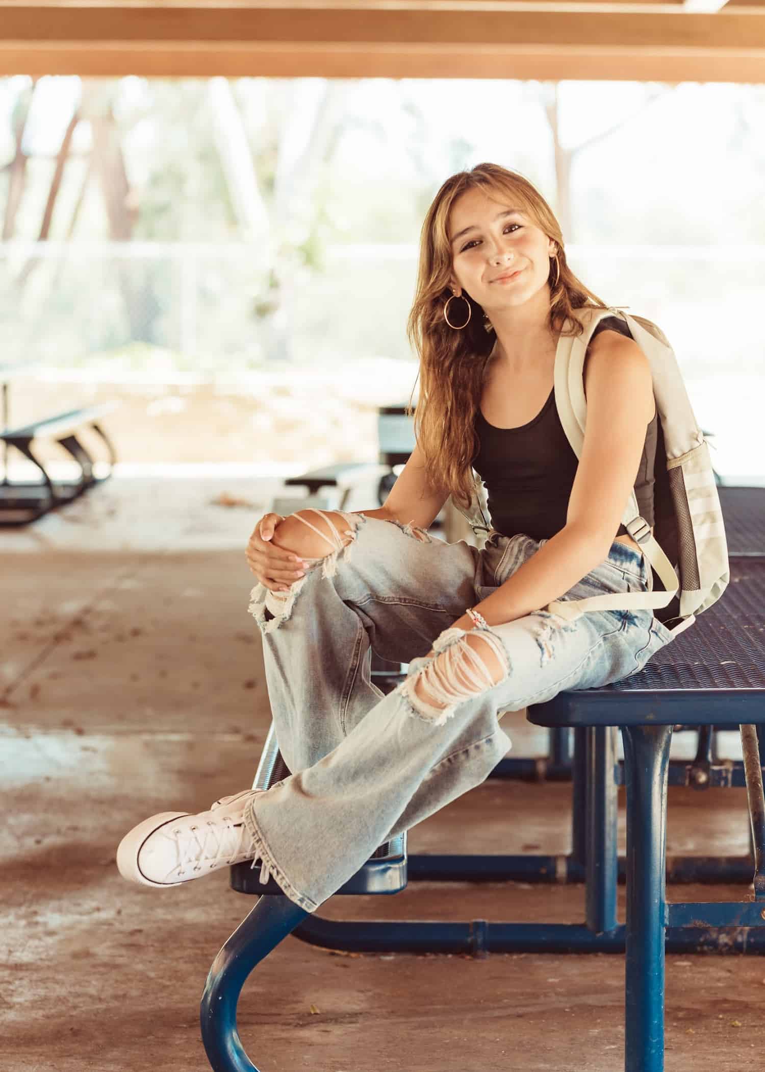 A pretty teen actress with long brown hair wearing ripped jeans and a black tank top. She is sitting on top of a picnic table smiling. 