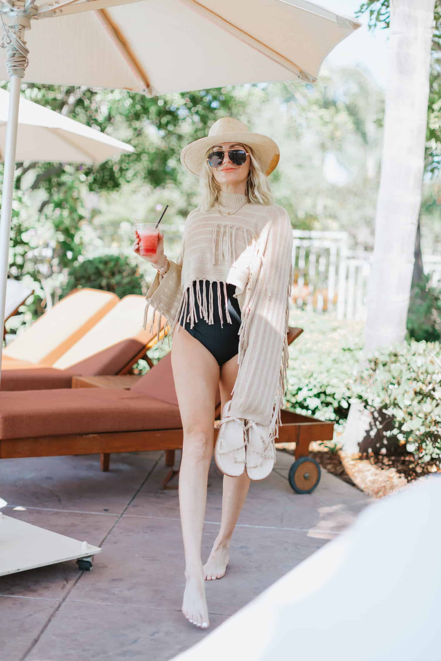 Attractive woman by the pool wearing a large brimmed hat, a  black swimsuit and a beige fringed coverup. Her skin has a healthy summer glow from wearing the best bronzer with sunscreen.