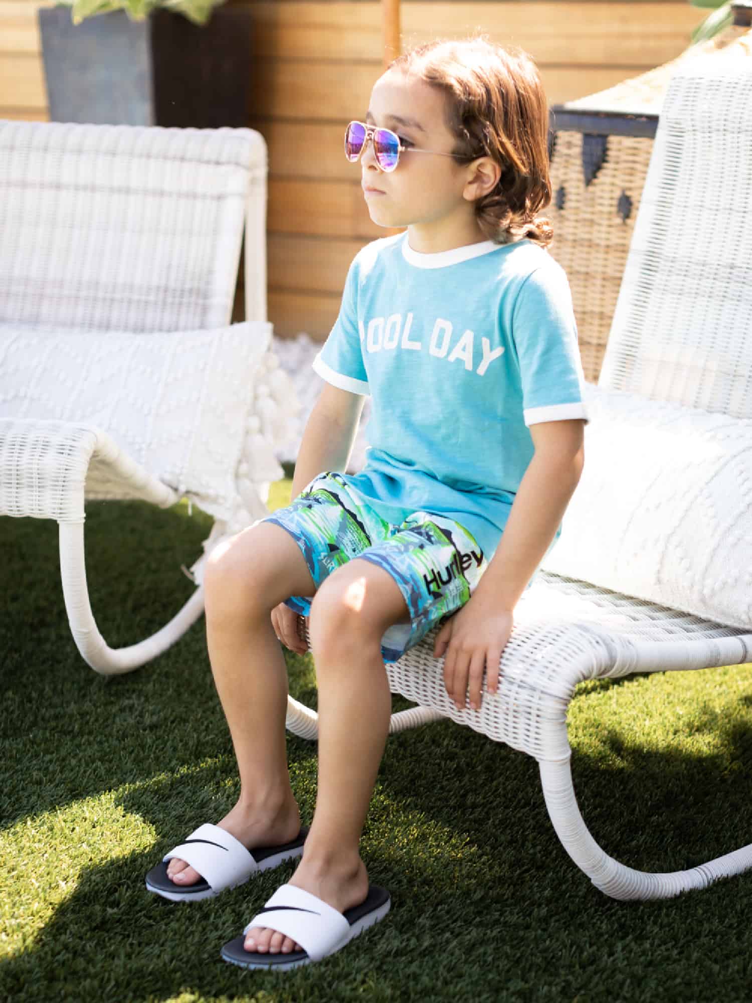 An adorable young boy is sitting on a pool chair. He is wearing blue print swim trunks from KOHL'S and a blue t-shirt. He's wearing sunglasses to shield him from the bright sun.