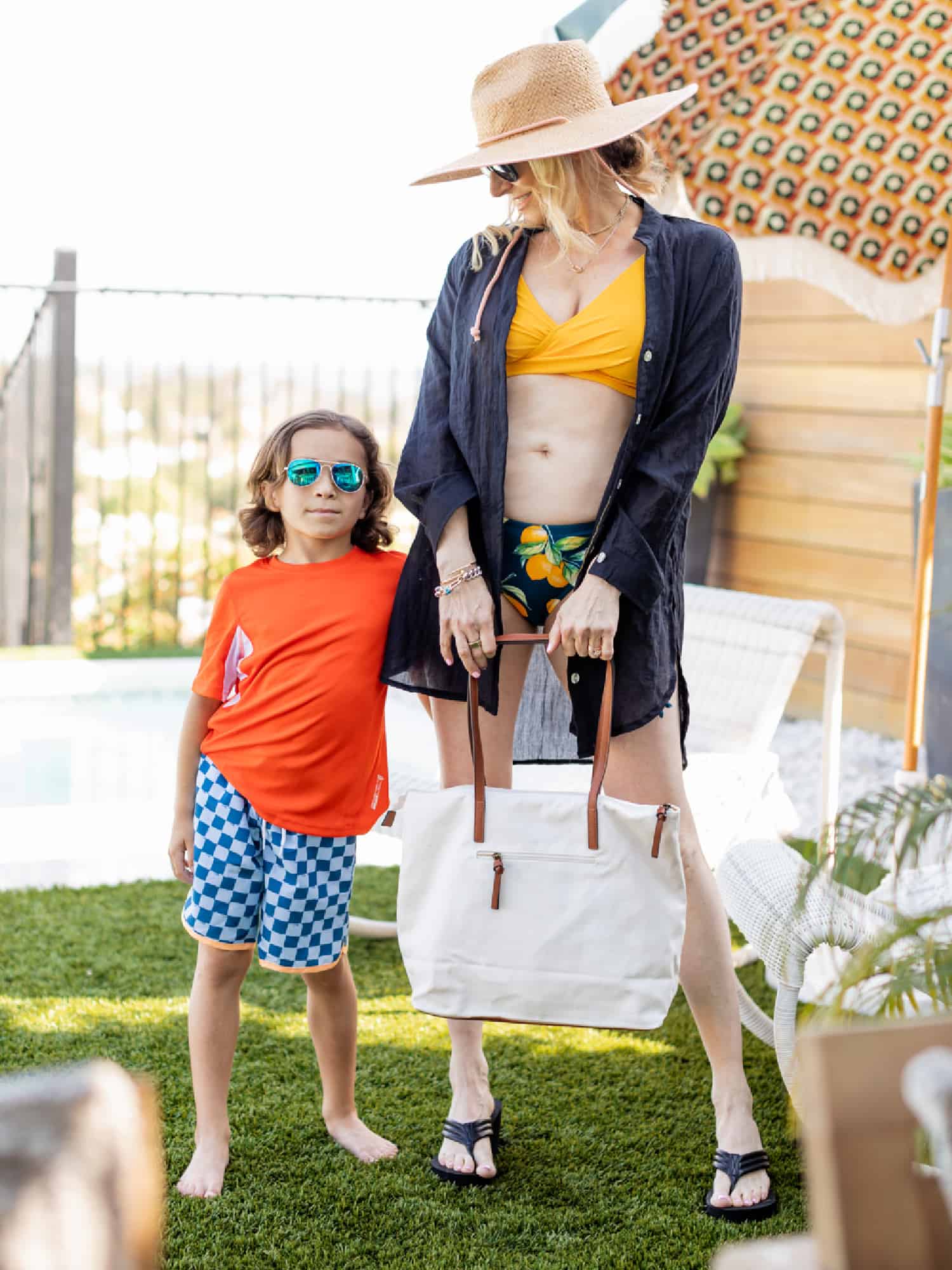 A happy mom and young son ready for a pool day. The mother is wearing a two-piece swimsuit with a bright yellow top and colorful bottom. She is accessorized with a blue long-sleeved beach cover-up, a large brimmed hat and a beach tote. The young boy is wearing colorful blue and white check swim bottoms and an orange t-shirt from KOHL'S.
