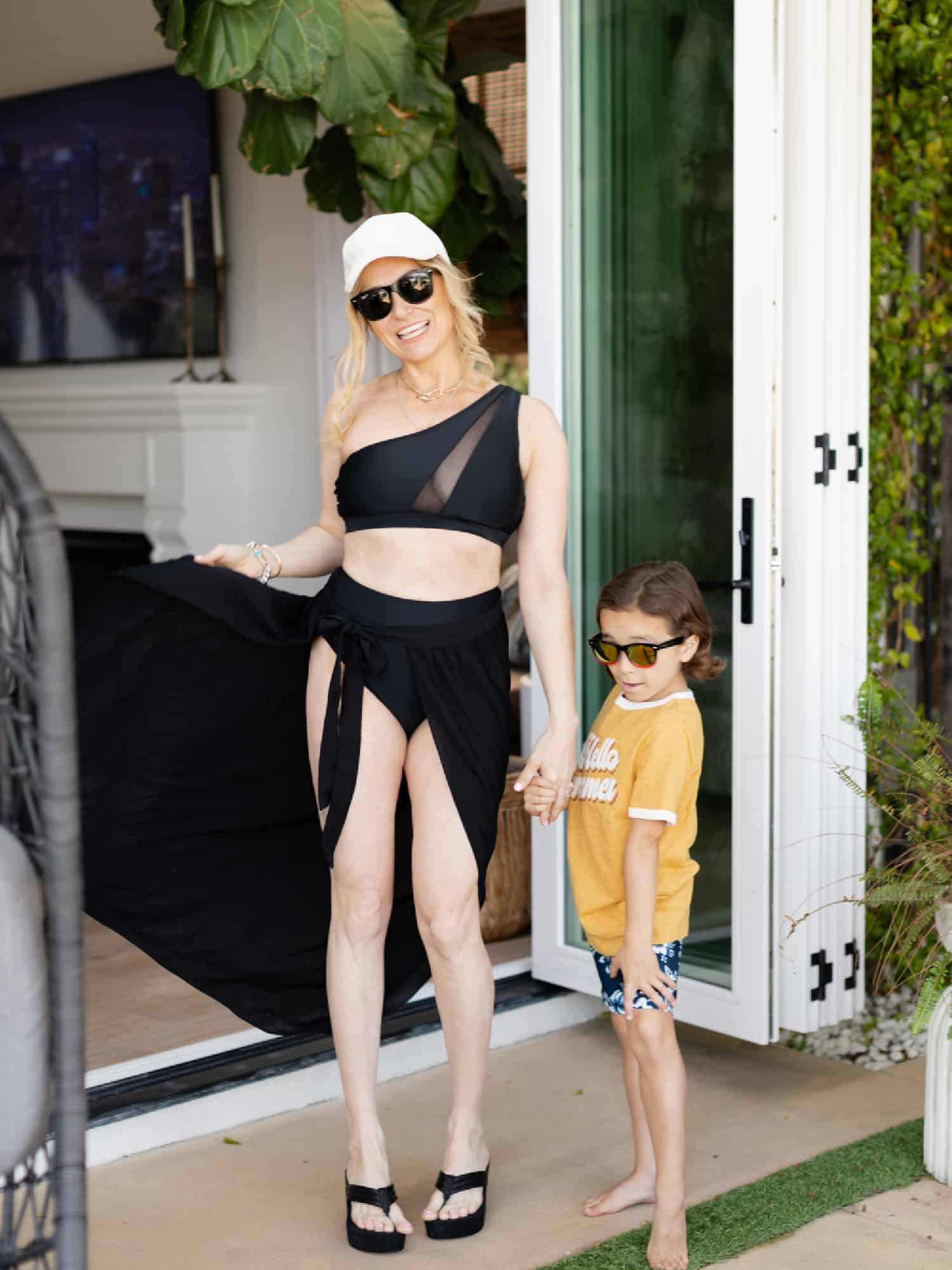Mother and young son wearing swimsuits. The mom is wearing a black two-piece with a black wrap skirt. She is also wearing a white ball cap and sun glasses. The boy is holding her hand and striking a cute pose. He's wearing blue swim trunks with a yellow t-shirt from KOHL'S.