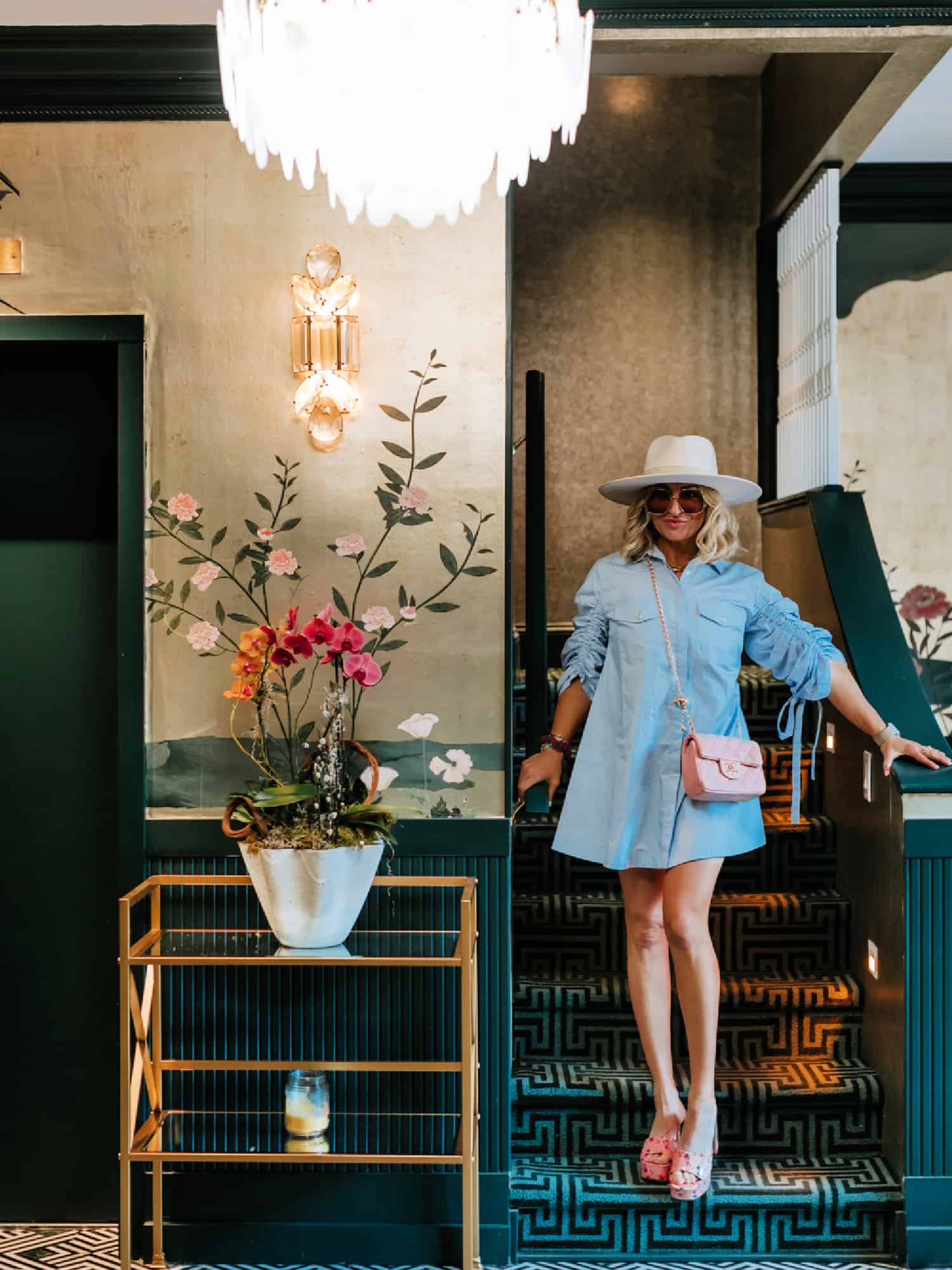 A woman standing on a step next to beautiful floral wallpaper, and a planter on a gold and glass table. She is wearing a light blue shirt-dress and a cream colored hat.