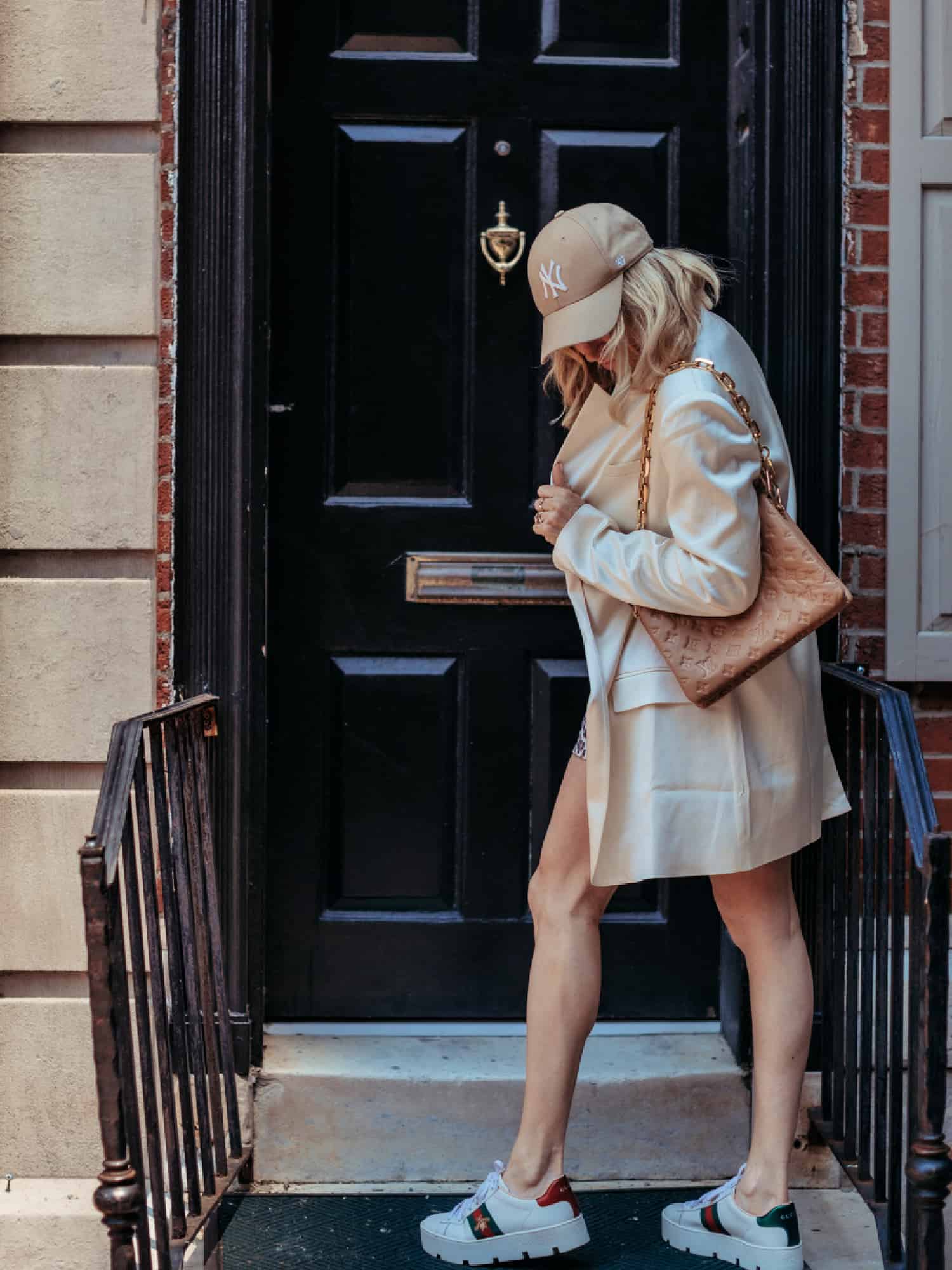 A woman standing in front of a black door while she looks down at her feet. She is wearing an oversized cream-colored blazer and a tan ballcap. 