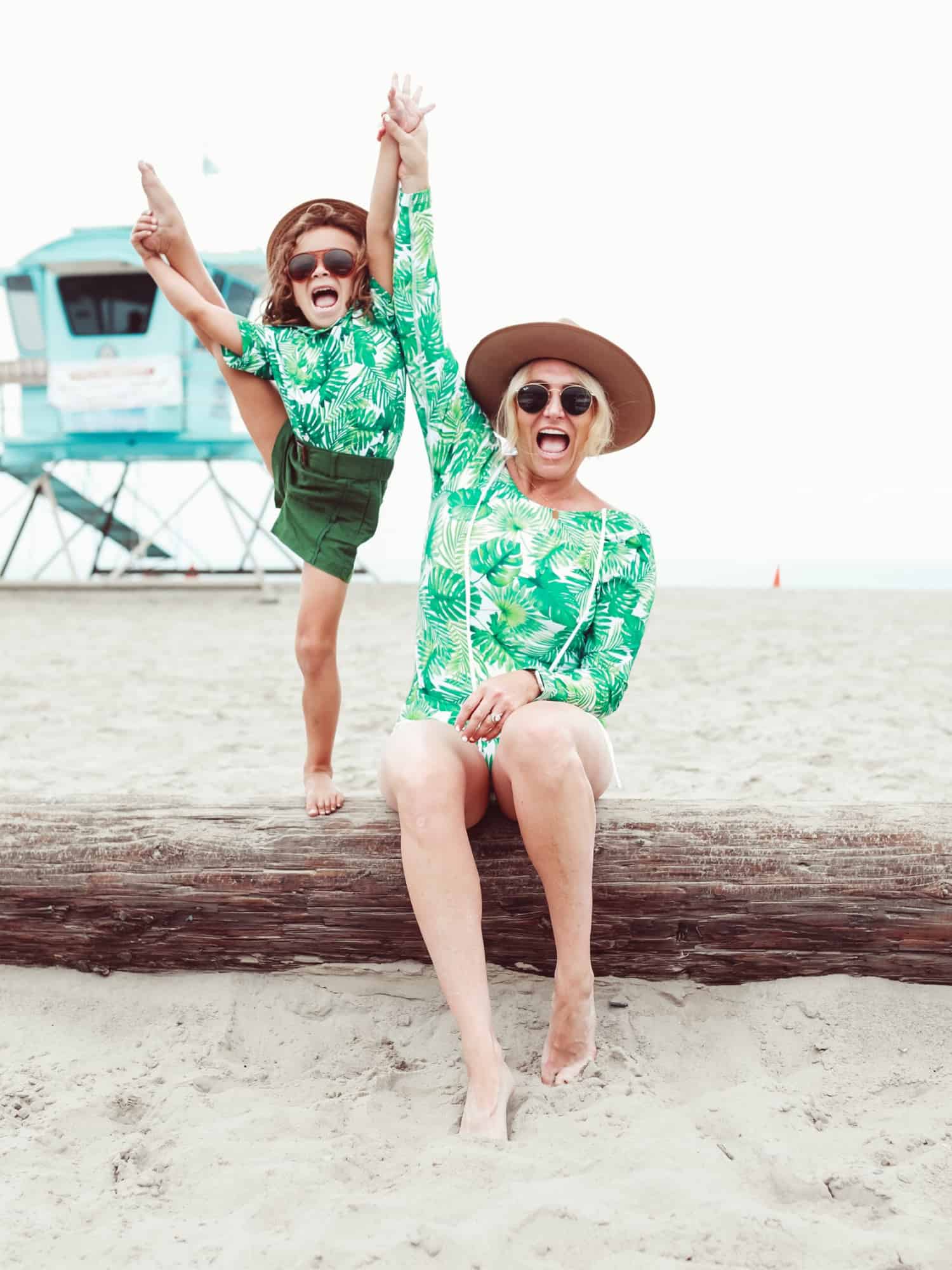 A woman wearing a green and white long-sleeved swimsuit with SPF protection. She is also staying protected from the sun by large sunglasses and a wide-brimmed hat. She is holding hands with a young boy wearing a similar swimsuit. They both appear to be having a fun time on the beach.