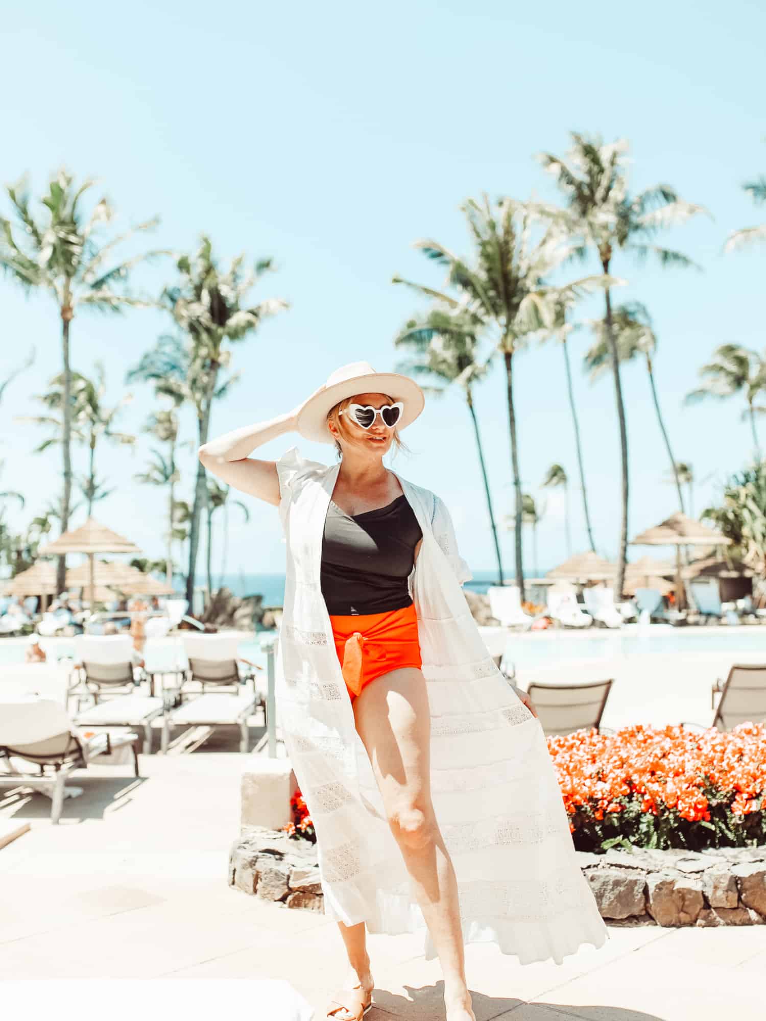 A woman wearing an orange and black swimsuit with a long white coverup. She has on white heart-shaped sunglasses and a large-brimmed hat for sun protection.