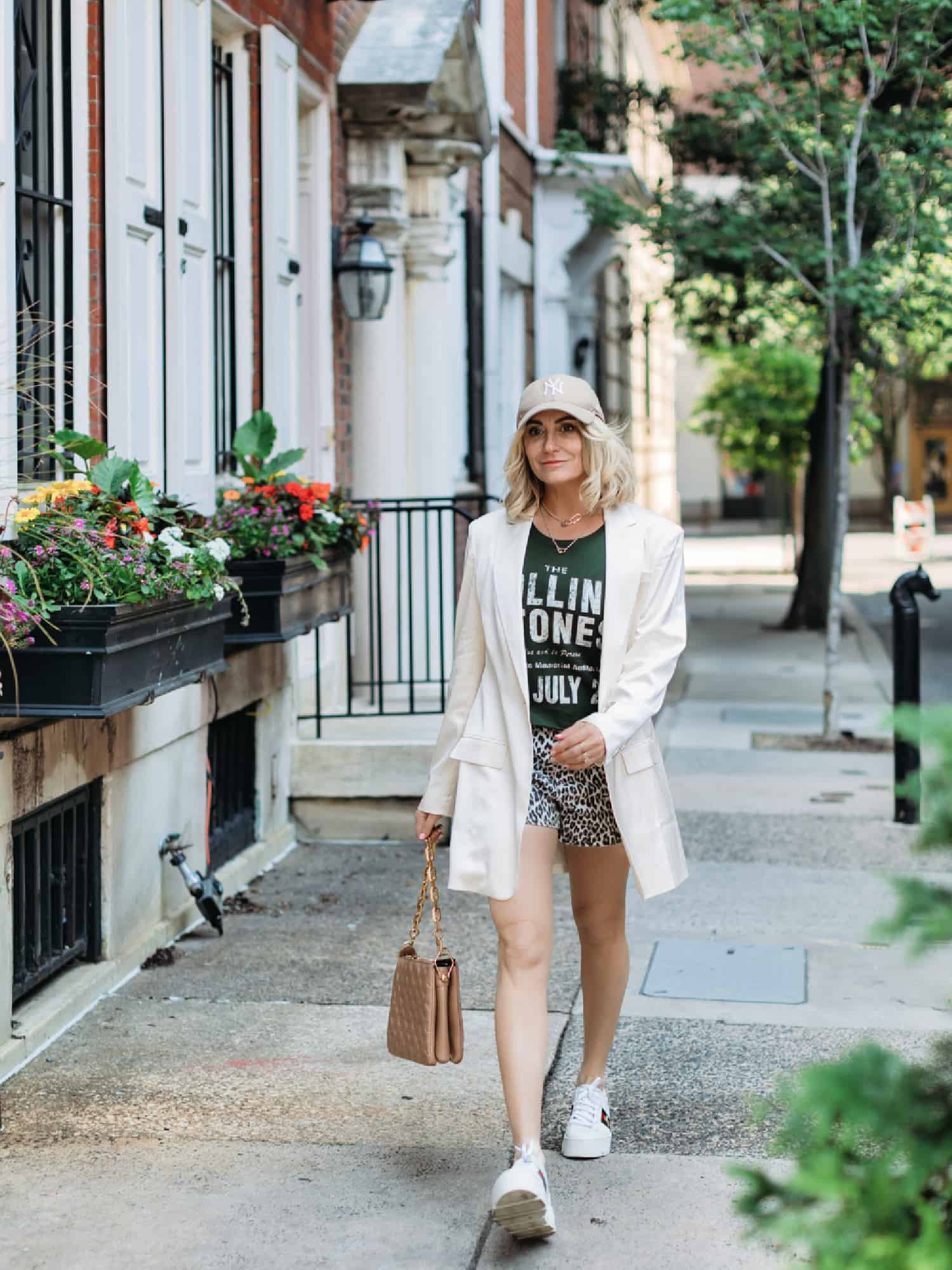 A woman looking summer chic, while she walks the city streets. She is wearing animal print shorts, a green graphic tee, and an oversized cream blazer. She is also wearing a cream-colored ball cap, and Gucci sneakers, while carrying a brown Gucci purse.