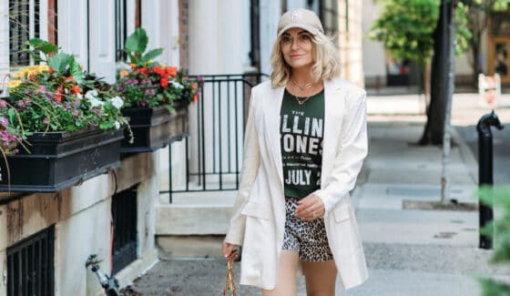 A stylish woman walking the city streets in a pair of animal print shorts, a green graphic tee, and oversized cream-colored blazer. She is also wearing a tan ball cap, white Gucci Sneakers and is holding a tan Gucci handbag.