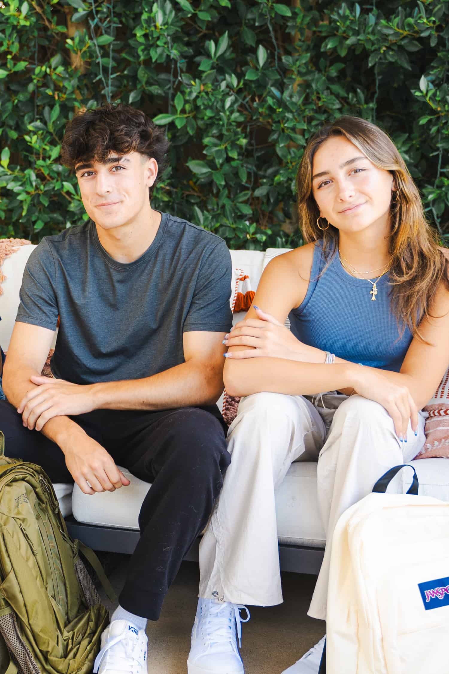 Attractive teenage boy and girl siblings sitting together on an outdoor sofa and discussing academic goals for high school. Two school backpacks sit on the ground in front of them. 