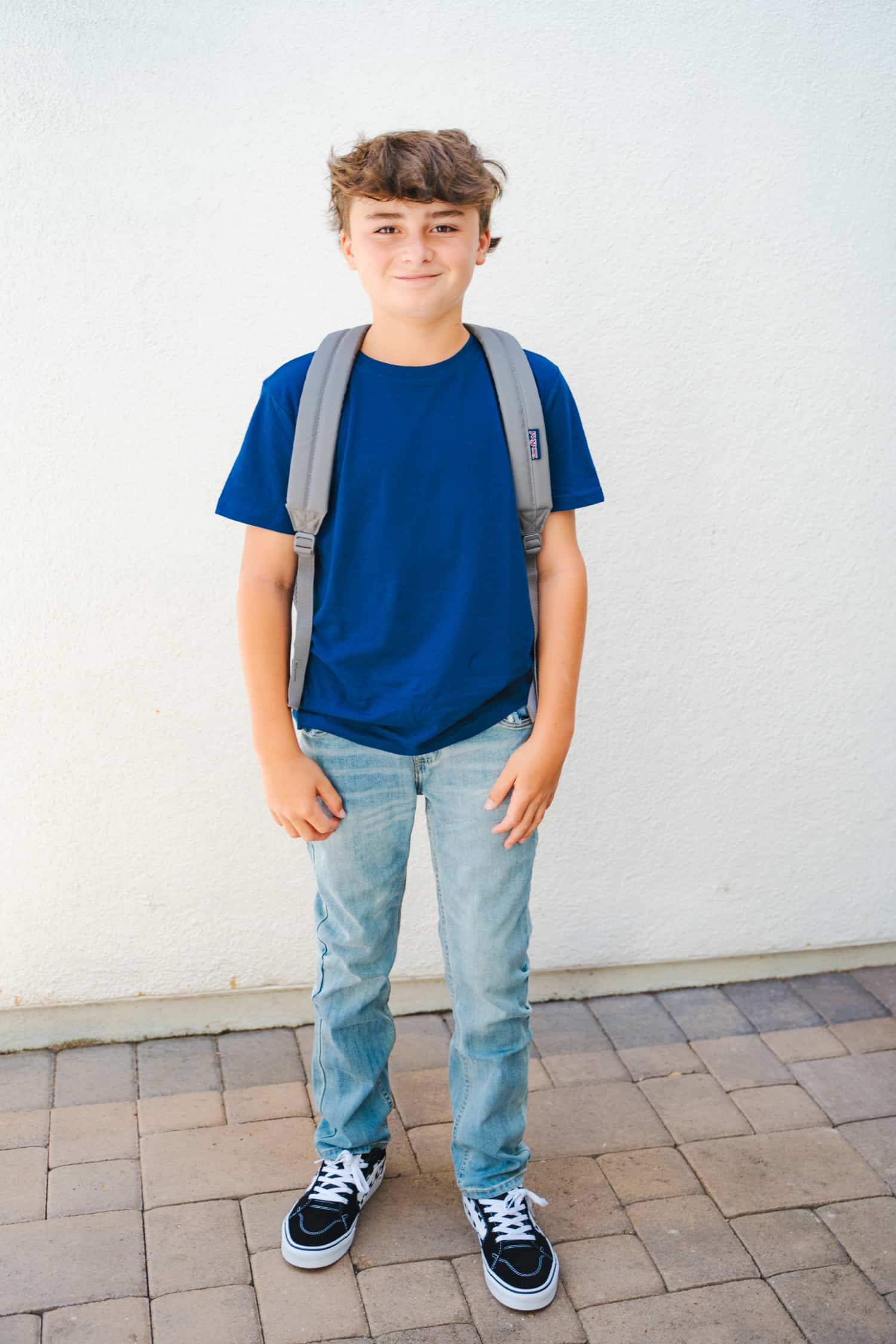 Adorable, smiling tween boy wearing Levi jeans, a bright blue t-shirt, black and white check Vans sneakers, and a grey JanSport backpack after back-to-school shopping at Kohl's.