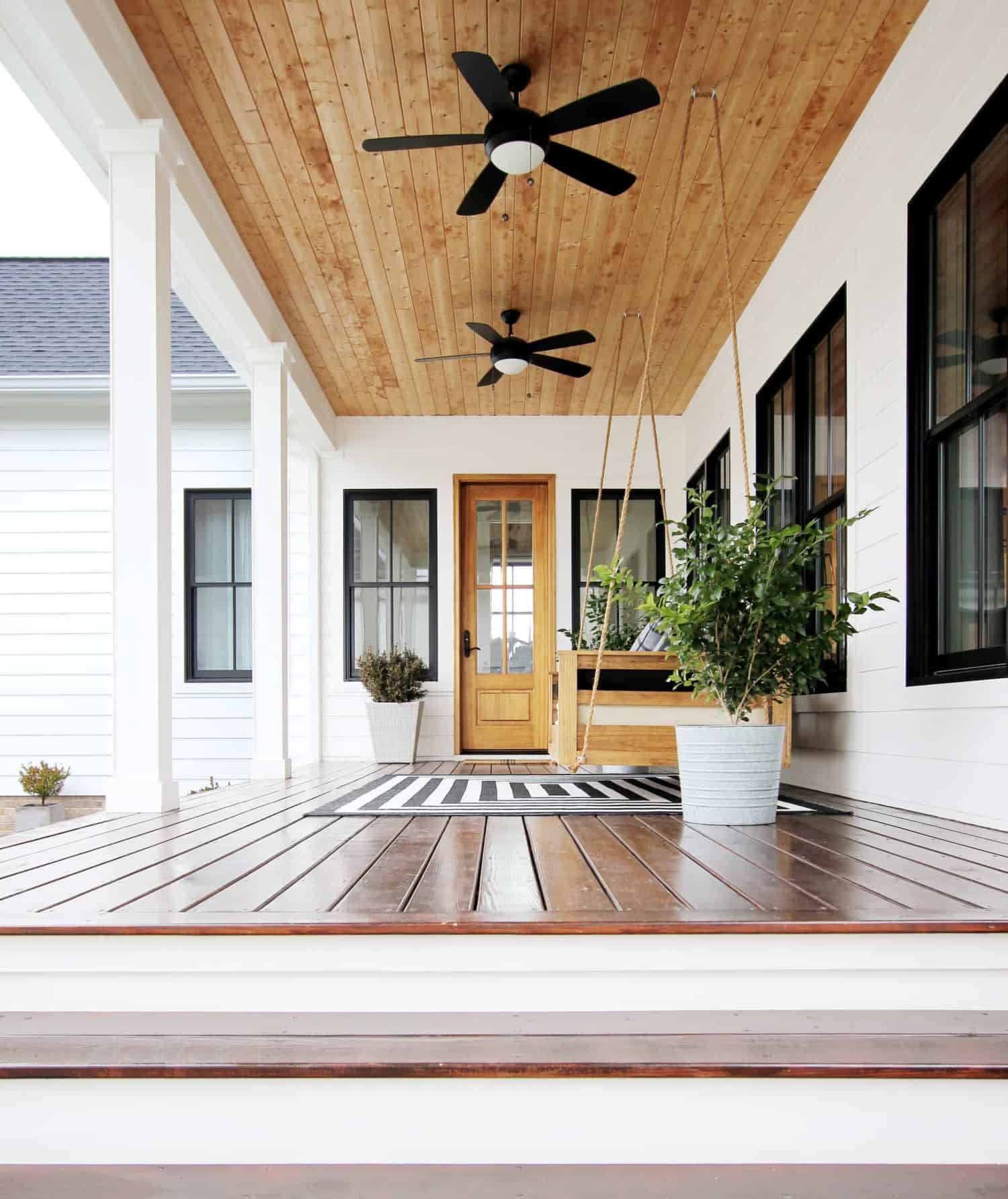Side view of a modern farmhouse porch. The porch is decorated with a simple black and white rug, plants in white planters and a large porch swing.
