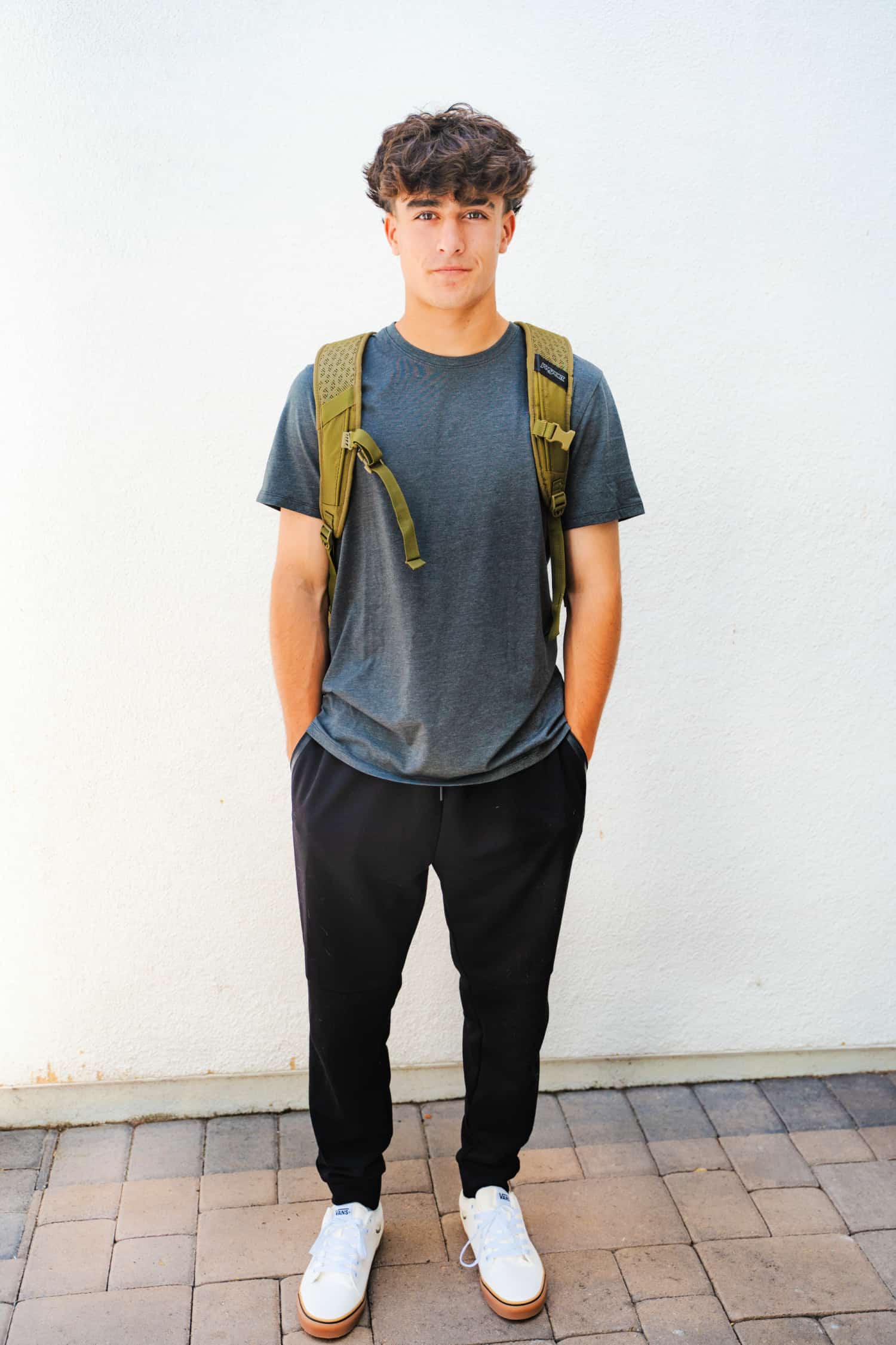 An attractive teen boy wearing back-to-school clothing from Kohl's consisting of black jogger pants, a blue t-shirt, white sneakers and a green backpack.